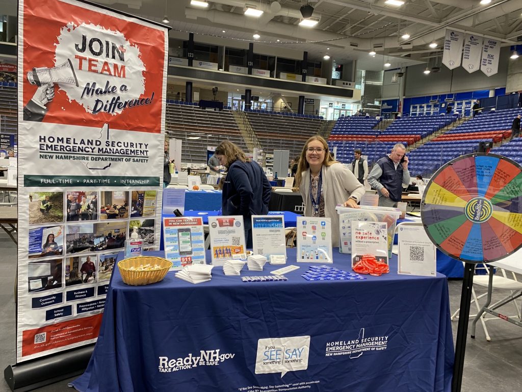 Hannah behind a table with a tablecloth and many items on top of it. To either side of the table is a large poster saying "Join the Team, make a difference"  and a spin-the-wheel on a stand. This is set up in an enormous gymnasium with red and blue seats.
