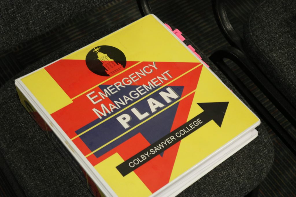 A Yellow, black and red binder design with arrows and white text reading "Emergency Management PLAN". A smaller arrow underneath reads "Colby-sawyer college". Everything is in all-caps, with "plan" in bold.