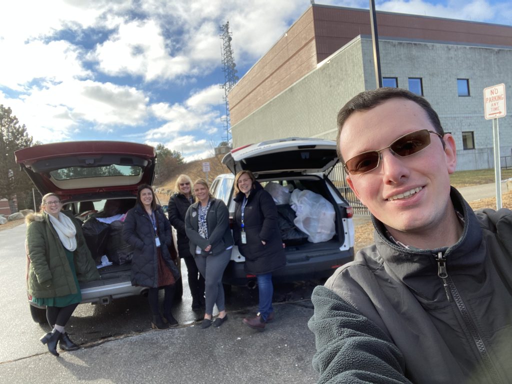 In a selfie done by Matt, multiple people pose in front of two large cars stuffed bags (which are in turn stuffed with gifts). The sun shines on a snow-free parking lot.