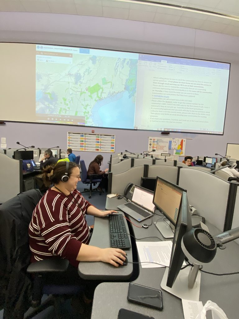On a large screen, a live map of the Northeast, and a blurry word document. Beneath them are people sitting at computers.