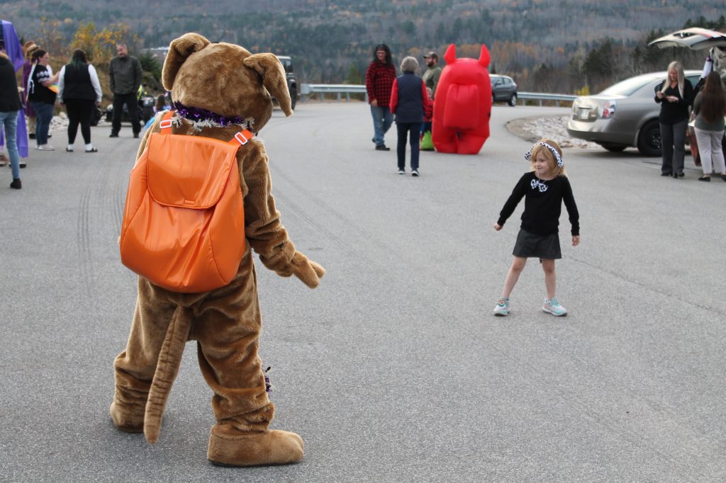 Ready the mascot stands surprised, 6 feet away from a very young girl in a defensive stance. She is glaring determined-ly at the mascot. Various DOC and trick or trreaters ar ein the backgground, parking lot with a glimpse of trees behind.