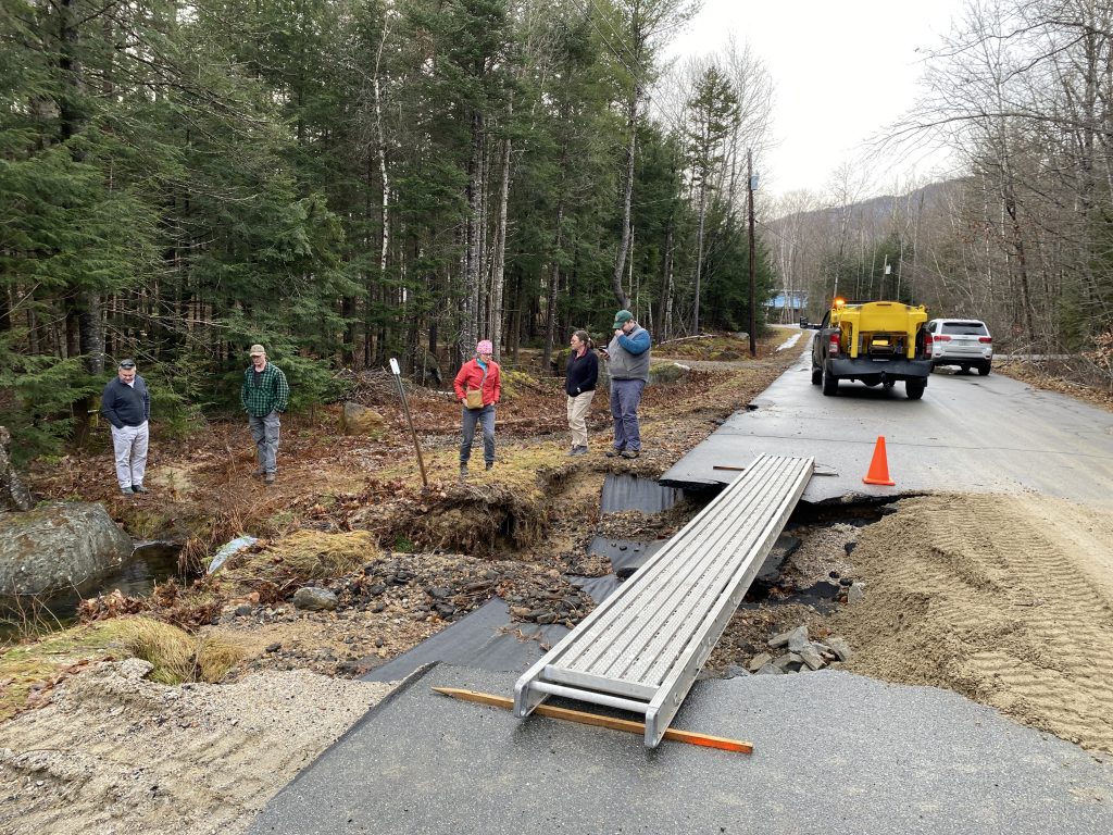 5 people stand near a collapsed section of roadway, in a wooded area. A metal device lies over the broken section of road, beyond which is a work truck and a car.