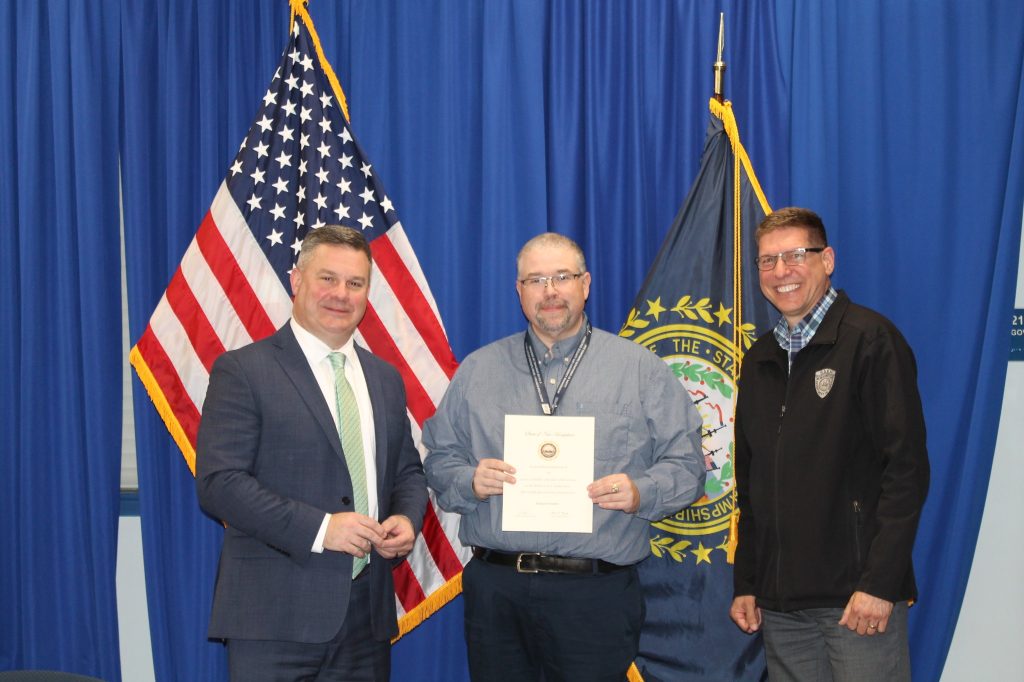 Robert B., Richard, and Robert C. stand in front of the NH state seal and a US flag. Richard holds up a congratulatory letter.