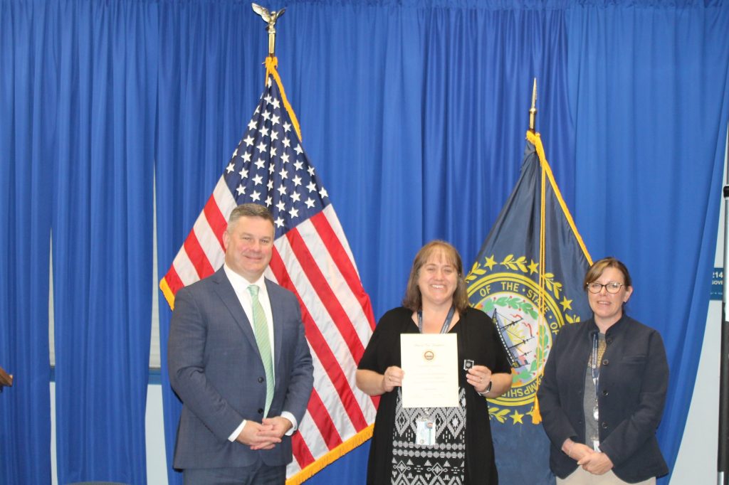 Robert B., Kelly, and Megan stand in front of the NH state seal and a US flag. Kelly holds up a congratulatory letter.