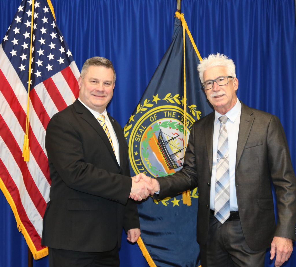 A photo of H S E M Director Robert Buxton and D o I T Commissioner Denis Goulet both wearing dark suits and shaking hands in front of the U S and New Hampshire state flags.