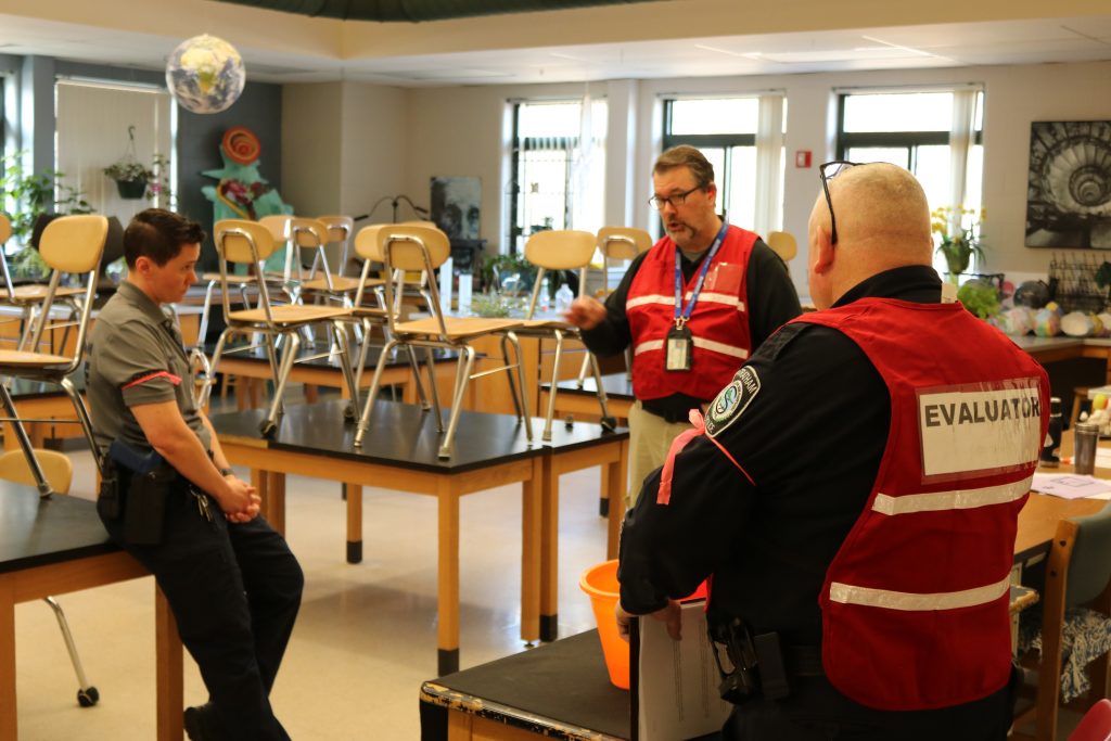 A photo of police officers and an HSEM employee talking in a school classroom filled with desks with chairs on top of the desks.