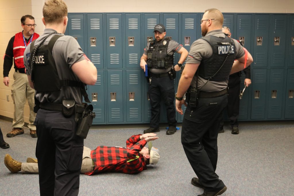 A photo of police officers and an HSEM employee standing in front of school lockers surrounding an actor lying on the floor with his hands in the air.