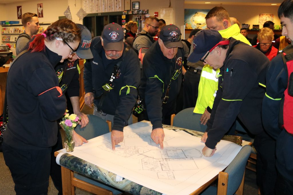 A photo of a group of first responders wearing their uniforms standing around a table that displays a schematic plan for the school.