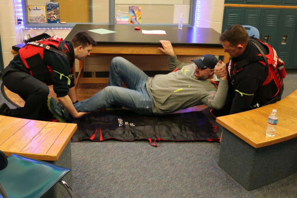 A photo of a man in a sweatshirt and jeans laying on the floor in a school classroom, being assisted by two first responders wearing medical vests.