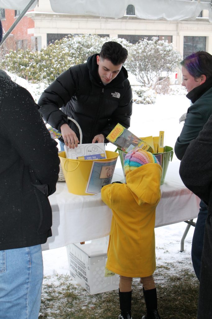 a small child in a yellow coat stands at a table with yellow buckets of goodies. Three people in black sweaters look on, framing the image.