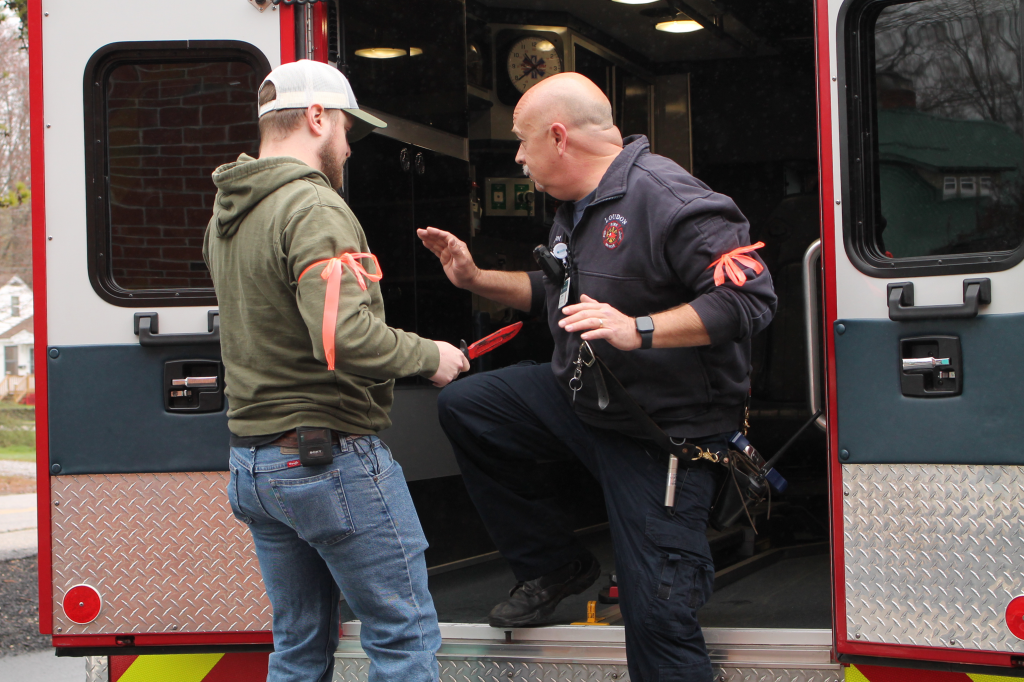 A man in a green sweatshirt and baseball cap holds an EMT at (rubber) knife-point. They are both wearing bright orange bands around their bicep.