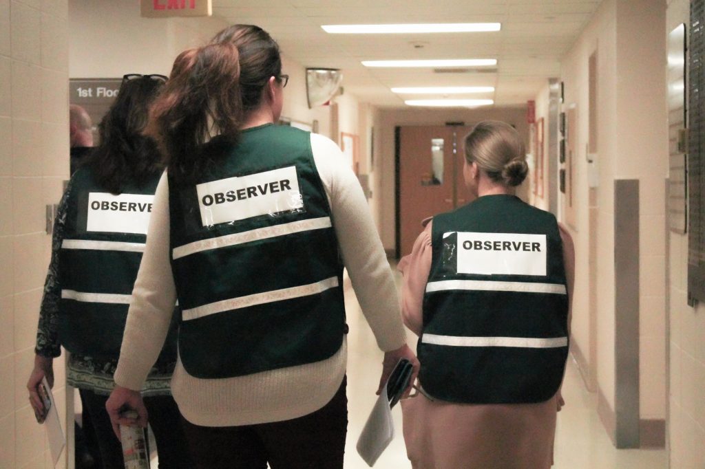 three women in green “observer” vests walking down a hallway