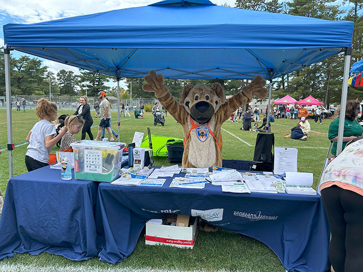 Ready the Prepared Puppy stands behind a table with his arms outstretched in the air. On the table are emergency preparedness materials.