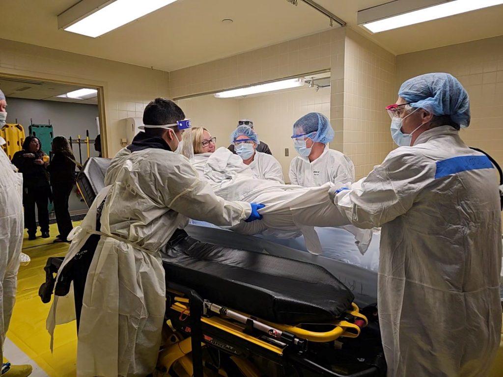 A woman lays on a gurney while a group of first responders in protective gowns, masks and wearing eye protection move her down a hallway in a hospital.