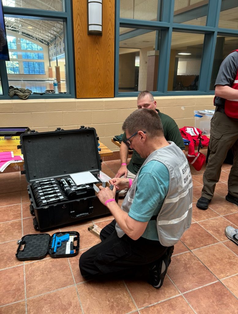 HSEM staff members look over equipment while sitting on the floor in front of windows leading into the Exeter High School cafeteria.